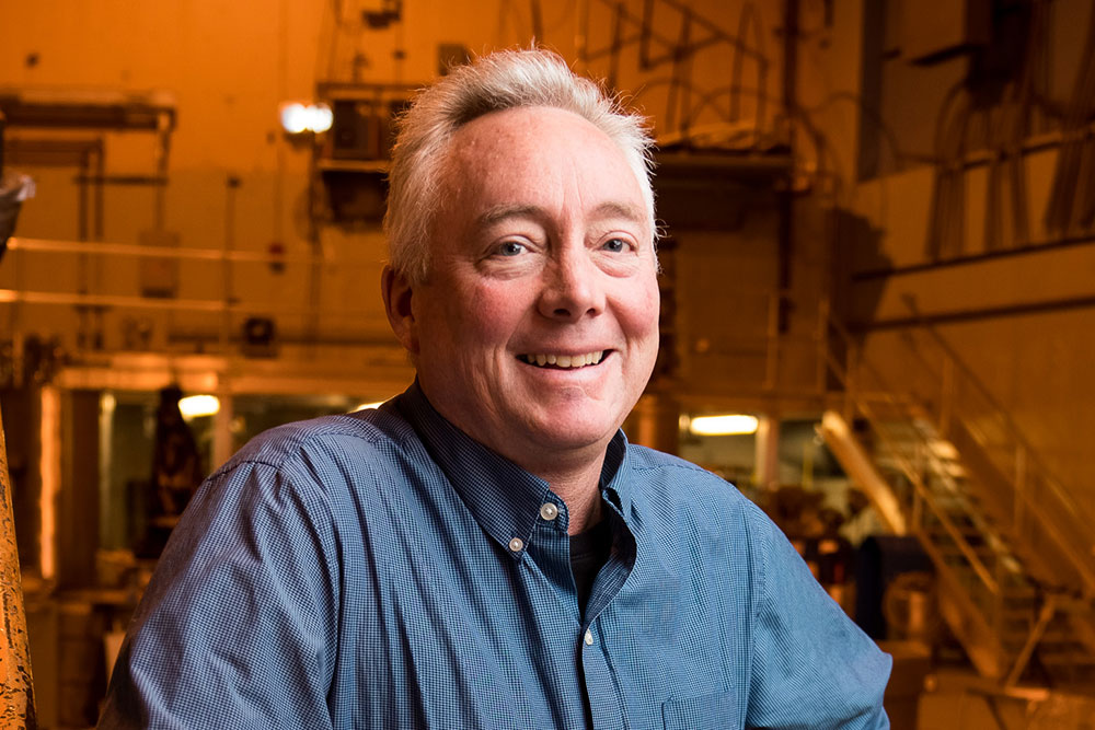 Dennis Whyte standing in large lab space at MIT with brick walls, staircase and balcony in background