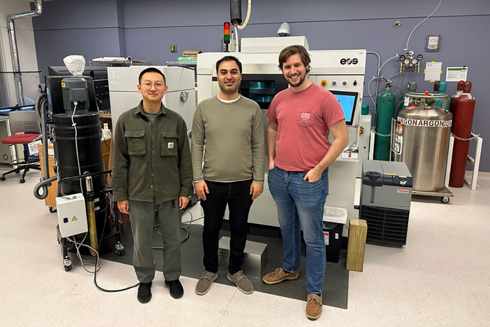 Three male students standing in a row in front of a 3D printer inside a lab, MIT