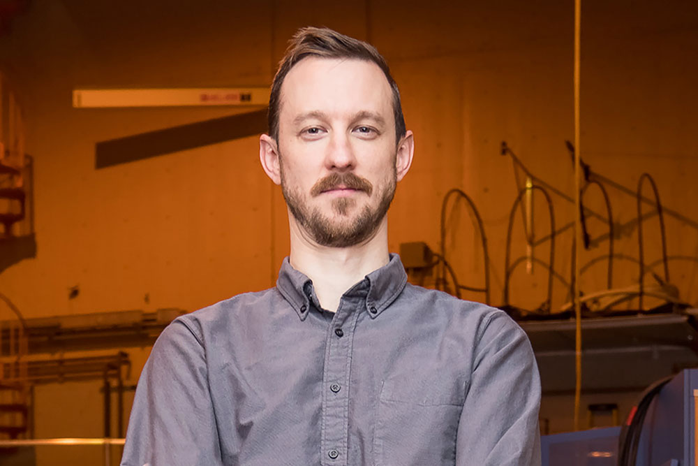 headshot of Zach Hartwig in the center of frame photographed in a lab space with orange lighting