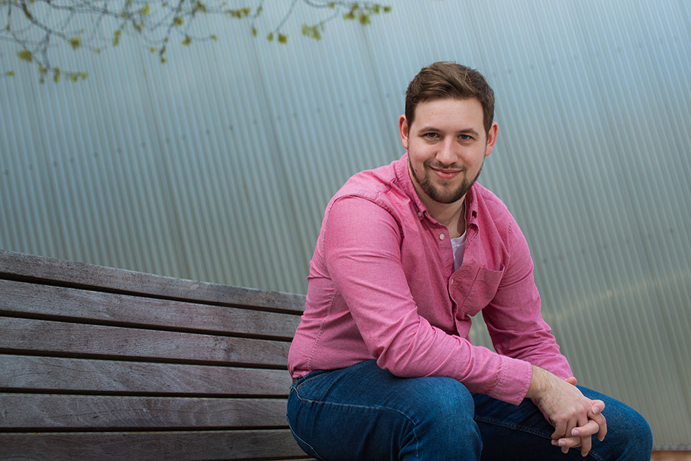 male student in red checked tshirt and jeans seated outdoors on park bench leaning forward with arms on knees with corrugated metal wall in background, MIT