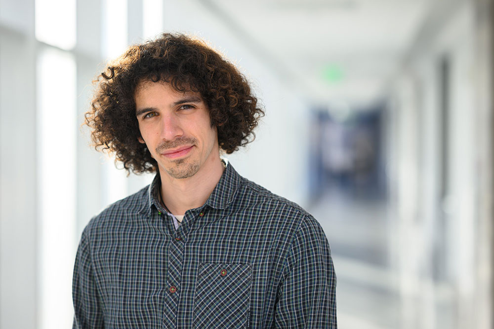 Graduate student, Eli Sanchez standing in a out of focus naturally lit hallway, in the left side of the photograph, MIT