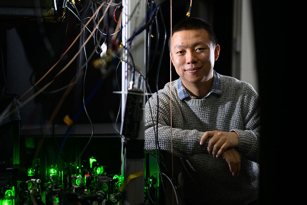 Male postdoc, Guoqing Wang, in lab leaning on his right arm; green laser instrumentation set-up in the foreground to the left and wires hanging above, MIT