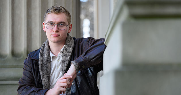 Graduate student, Liam Hines, seated outdoors with concrete architectural columns in the background and to the right